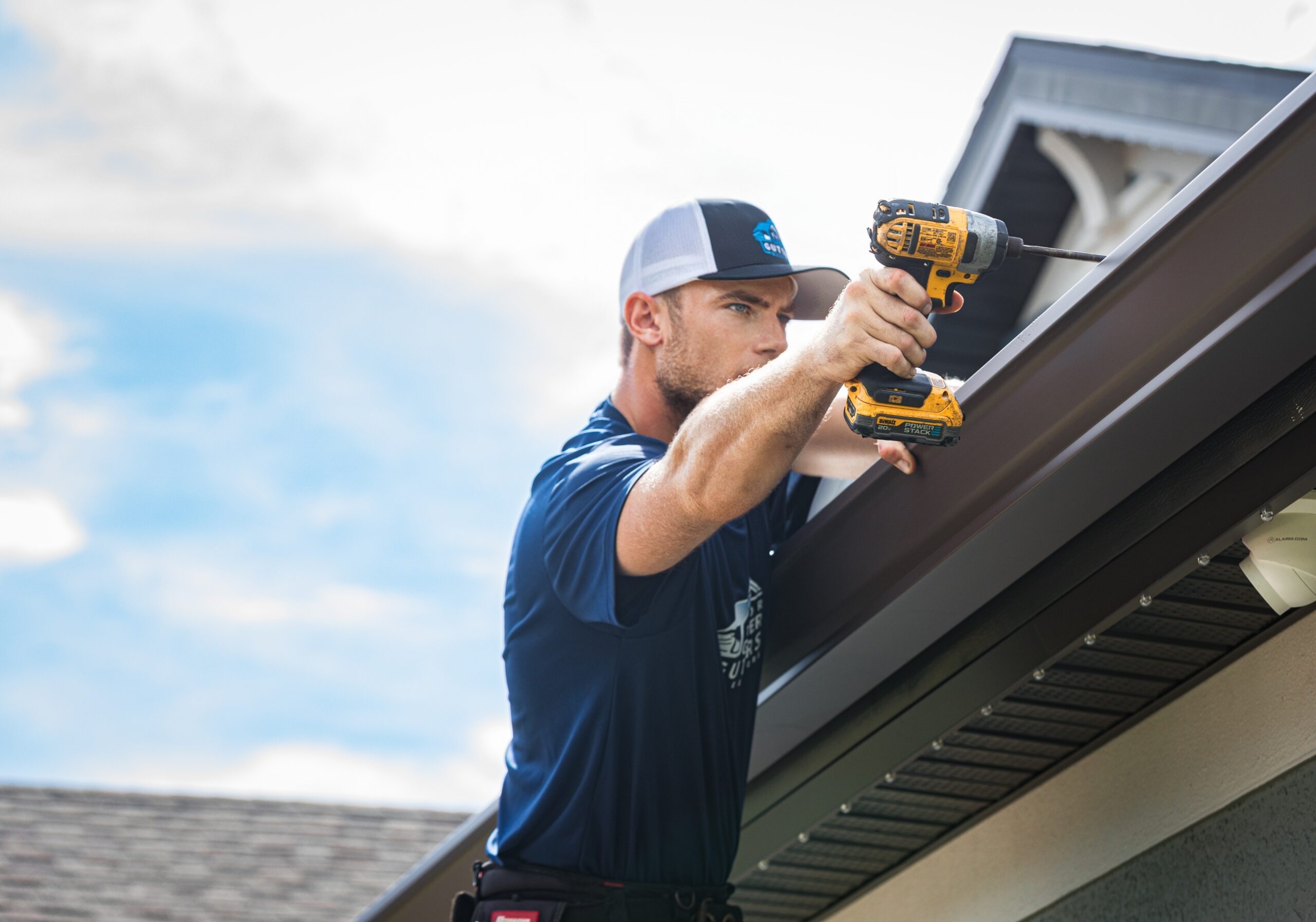 A technician using a yellow power drill to install dark brown gutters on a residential roof